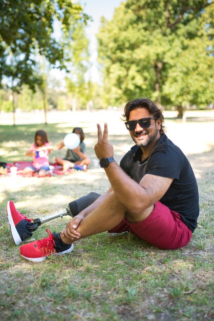 Retrato de hombre contenido con discapacidad en picnic en el parque. Hombre de cabello oscuro con gafas de sol posando, sentado en la hierba, mostrando el signo de la victoria. Niños en segundo plano. Discapacidad, familia, concepto de amor