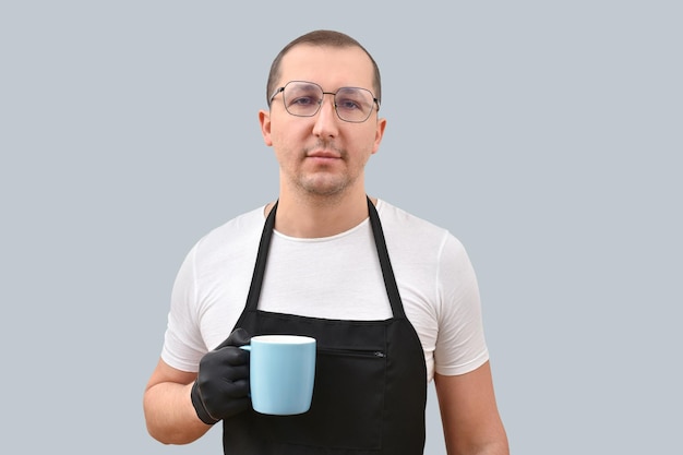 Retrato de un hombre en un delantal con una taza en las manos mirando a la cámara sobre un fondo gris