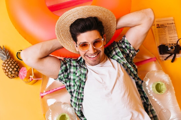 Foto retrato de un hombre descansando sobre un colchón inflable hombre con gafas de sol naranjas y sombrero de paja está acostado entre una cámara retro de cóctel y un periódico