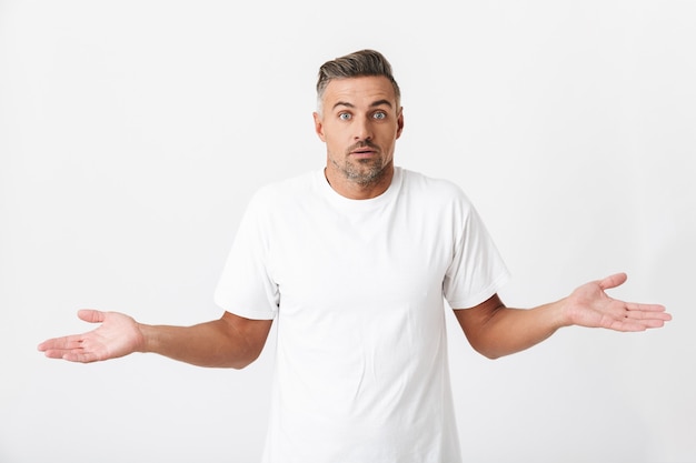 Retrato de hombre desconcertado de 30 años con cerdas vistiendo camiseta casual levantando las manos aisladas en blanco