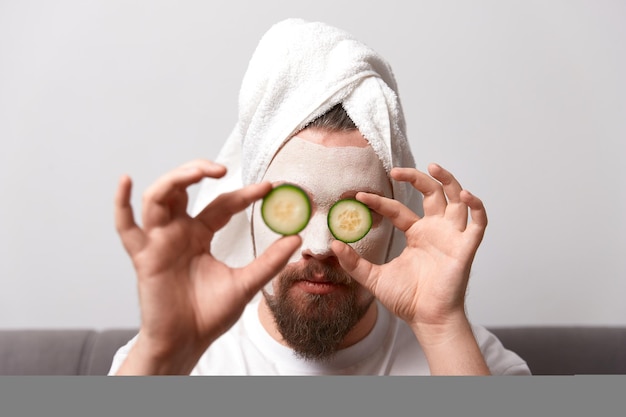 Retrato de un hombre divertido y feliz con camiseta blanca aplicando rodajas de pepino sobre una máscara de arcilla en la cara. Procedimiento de autocuidado en el spa matutino. Concepto de tratamiento de belleza