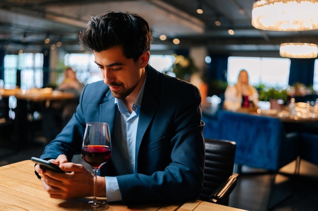 Retrato de un hombre elegante y apuesto que usa traje de moda usando un teléfono inteligente charlando con amigos y bebiendo vino tinto sentado en la mesa en el restaurante