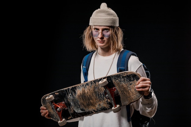 Retrato de un hombre elegante que sostiene una patineta en el estudio. Primer plano de un skater sonriente posando sobre un fondo negro.