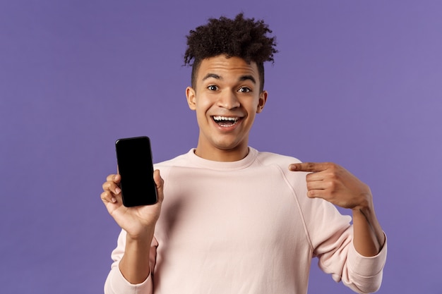 Retrato de un hombre emocionado, feliz y sonriente que habla sobre una nueva aplicación o característica del juego, apuntando a la pantalla del teléfono inteligente, hablando sobre un teléfono móvil con expresión alegre y divertida