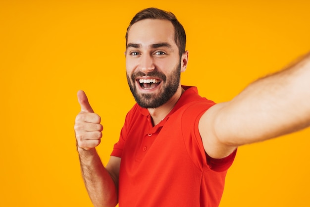 Retrato de hombre encantado en camiseta roja riendo y mostrando el pulgar hacia arriba mientras toma una foto selfie aislada sobre amarillo