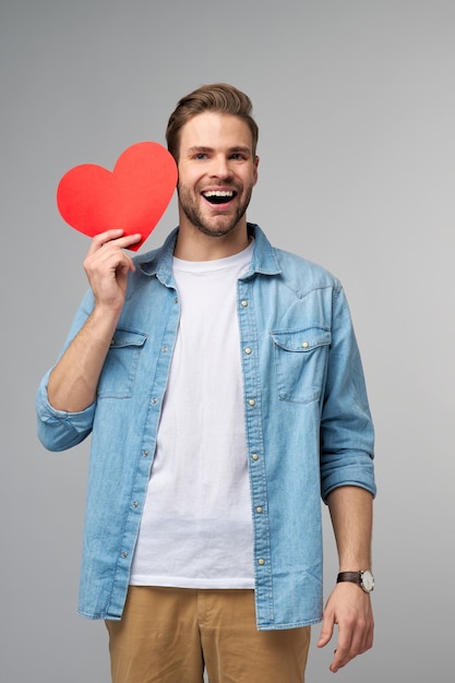 Retrato de hombre encantador mantenga la mano de papel en forma de corazón de la tarjeta use camisa de estilo casual aislado sobre gris