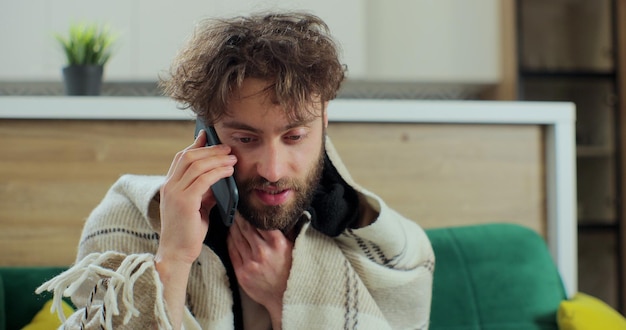 Retrato de un hombre enfermo sentado en la habitación y hablando por teléfono mientras se sienta en el sofá. Hombre enfermo tosiendo y teniendo dolor en el pecho en casa mientras habla por teléfono inteligente.