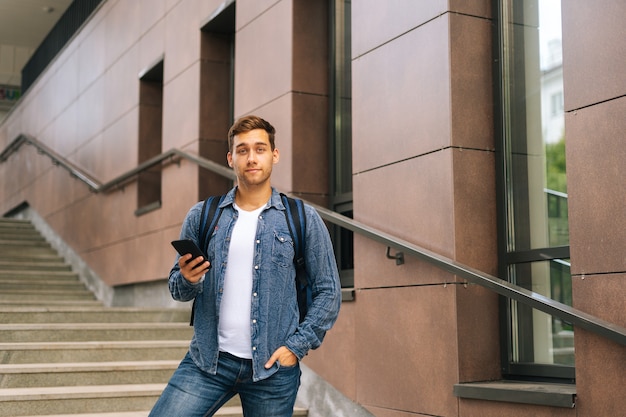 Retrato de hombre de entrega guapo con mochila termo grande sosteniendo usando teléfono móvil de pie en las escaleras del edificio de oficinas, mirando a cámara, fondo borroso, enfoque selectivo.