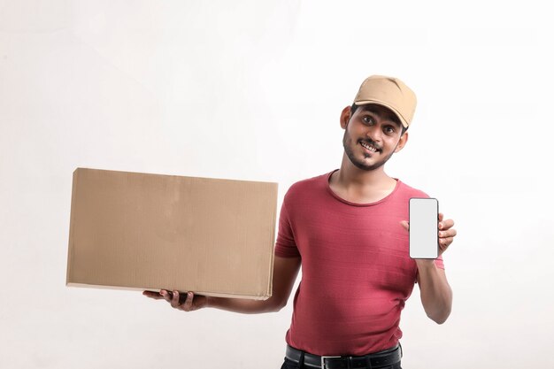 Retrato de un hombre de entrega joven feliz emocionado en la tapa que se coloca sobre el fondo blanco. Mirando la cámara que muestra la pantalla del teléfono móvil.