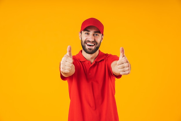 Retrato de hombre de entrega morena en camiseta roja y gorra sonriendo y mostrando el pulgar hacia arriba aislado sobre amarillo