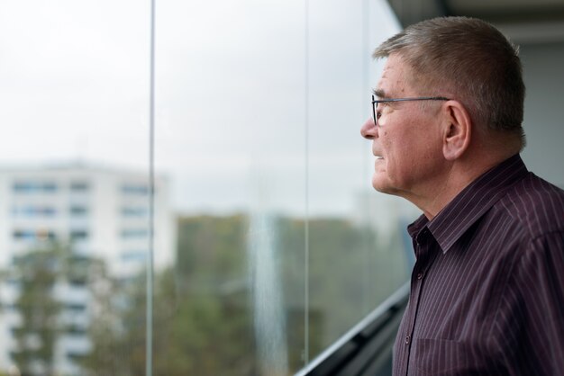 Retrato de hombre escandinavo senior con cabello gris con anteojos mientras mira por la ventana en el interior