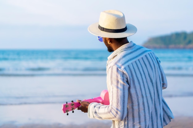 Retrato de hombre de etnia negra disfruta de las vacaciones de verano en la playa tocando el ukelele junto al mar