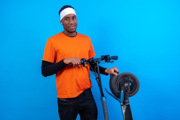 Retrato de un hombre de etnia negra con ropa naranja en un fondo azul con una patineta eléctrica