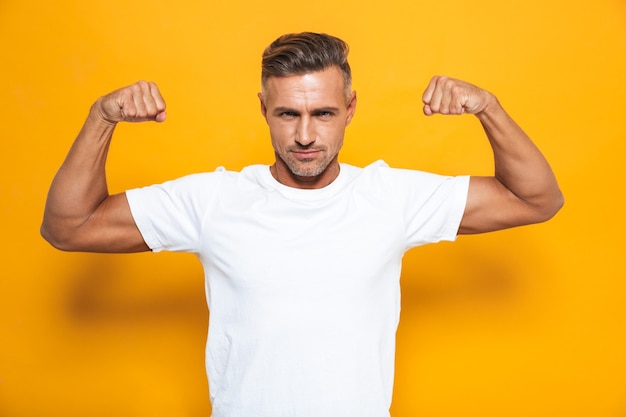 Retrato de hombre europeo de 30 años en camiseta blanca levantando las manos y mostrando bíceps aislado en amarillo