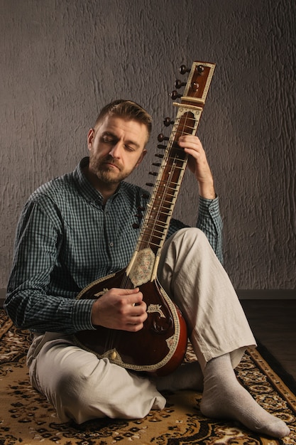 Retrato de un hombre europeo tocando el sitar sentado en la alfombra