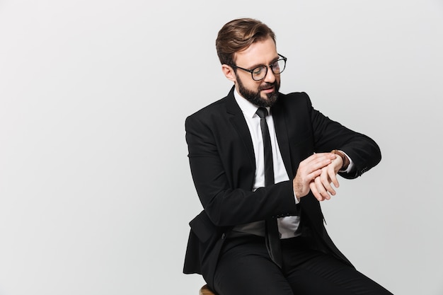 Retrato de hombre exitoso en traje y anteojos sentado en una silla y mirando el reloj de pulsera, aislado sobre la pared blanca