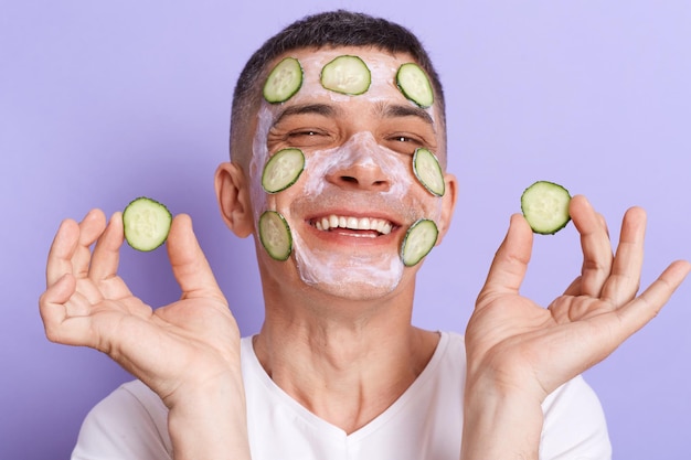 Retrato de un hombre extremadamente feliz y sonriente con un pantalones blanco que se aplica una máscara con rodajas de pepino en la mano y se las pone en la cara aislado sobre un fondo morado