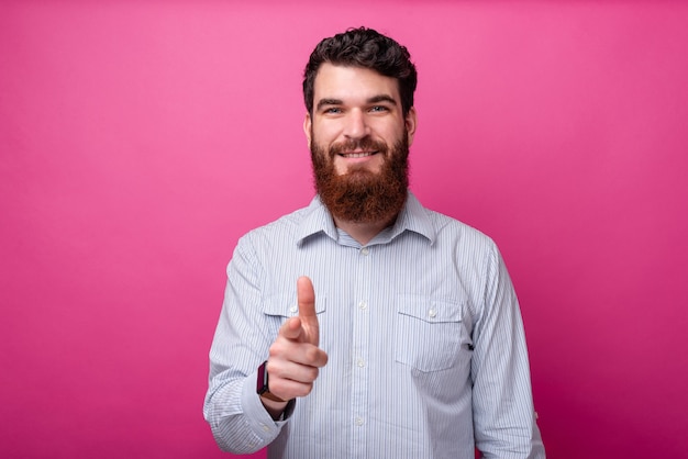 Retrato de hombre feliz con barba en casual apuntando a la cámara mientras está parado sobre fondo rosa