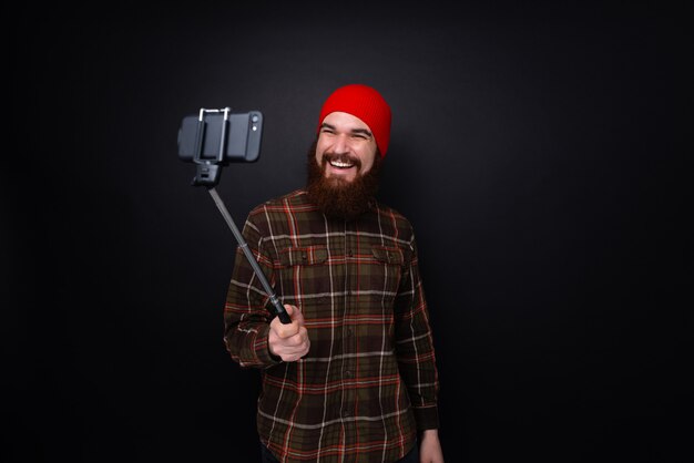 Retrato de un hombre feliz con barba haciendo selfie foto con palo aislado en una pared oscura