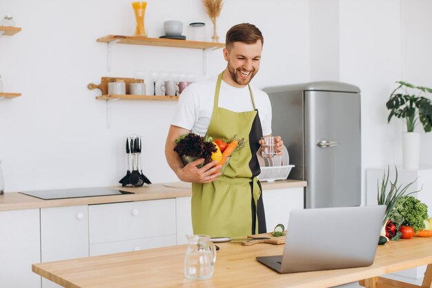 Retrato de hombre feliz en casa hombre cocinando ensalada de verduras mirando a la cámara y sonriendo rebanando verduras usando una computadora portátil para el entrenamiento de cocina en línea