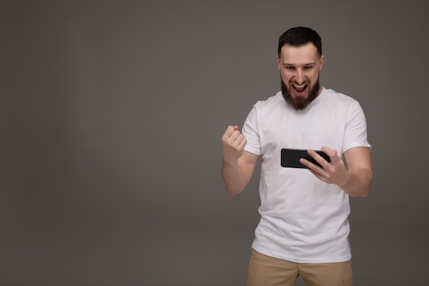 Retrato de un hombre feliz confía en sostener el teléfono móvil y celebrar el éxito aislado sobre fondo gris