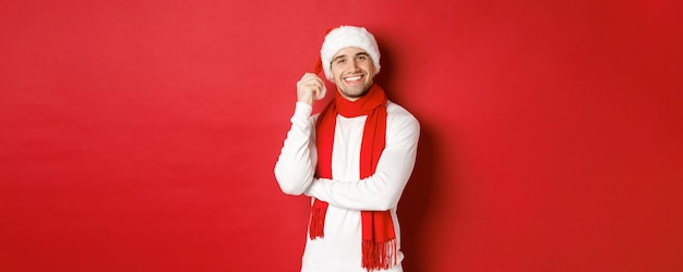 Retrato de hombre feliz disfrutando de las vacaciones de navidad con sombrero de santa y bufanda sonriendo alegremente de pie...