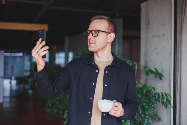 Retrato de un hombre feliz y exitoso con anteojos ópticos sosteniendo un teléfono móvil moderno y café en sus manos un blogger alegre con un teléfono inteligente digital posando en un café