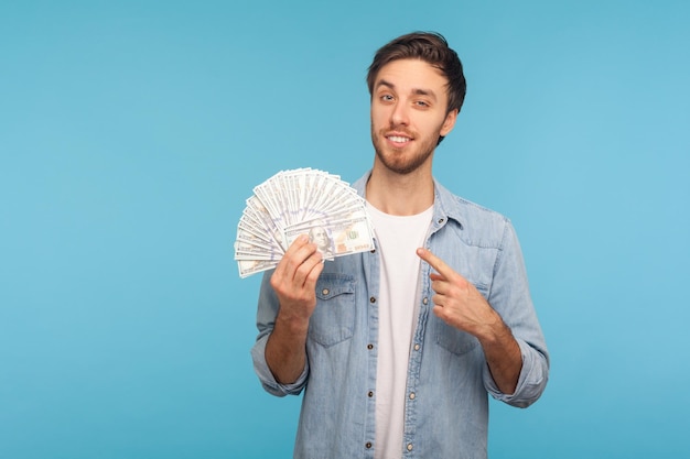 Retrato de un hombre feliz y guapo con camisa de mezclilla de trabajador que señala billetes de dólar y sonríe a la cámara alardeando de grandes ganancias de dinero regocijándose de la lotería gana una foto de estudio interior aislada en el fondo azul