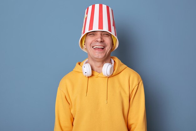 Retrato de un hombre feliz y rebosante de alegría con un gorro y una capucha amarilla informal posando contra una pared azul de pie con un cubo de palomitas de maíz en la cabeza riéndose a carcajadas divirtiéndose en el cine