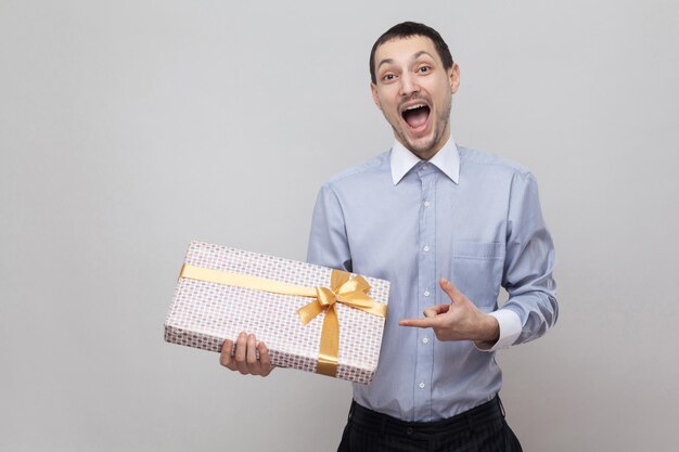 Retrato de hombre feliz satisfecho con camisa azul de pie y señalar con el dedo para presentar con lazo amarillo y boca abierta, mirando a la cámara. Interior, aislado, foto de estudio, fondo gris, espacio de copia