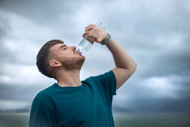 Retrato de un hombre feliz y sediento, un joven apuesto que está bebiendo agua mineral pura y fresca de una botella de plástico en el fondo natural del océano en un país tropical sosteniendo una botella en la mano
