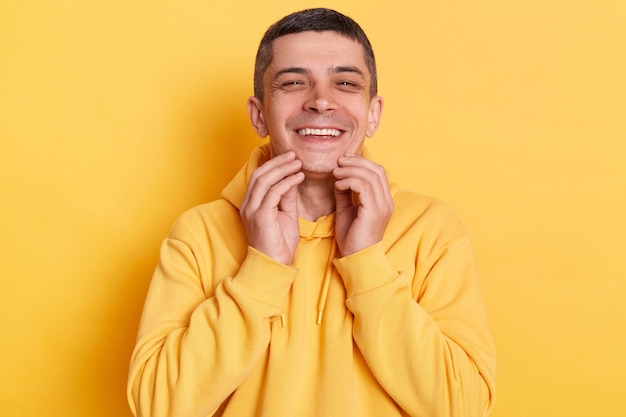 Retrato de un hombre feliz y sonriente con capucha de estilo informal posando aislado sobre fondo amarillo mirando la cámara con una sonrisa tocándose la barbilla