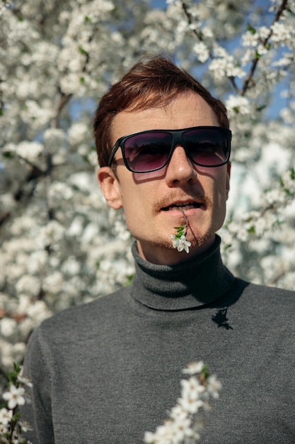 Retrato de hombre con flor en la boca mirando a la cámara en el jardín de primavera con árboles florecientes