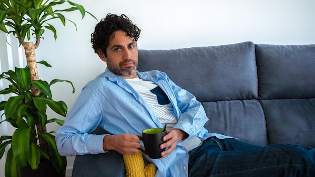 Retrato de hombre fresco y guapo bebiendo café expreso matutino y sentado en un sofá en el interior de la casa. Una persona está descansando y mirando hacia otro lado.