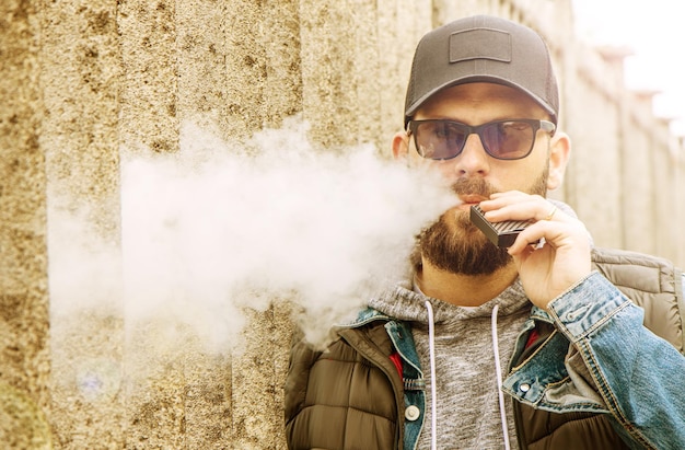 Foto retrato de un hombre con gafas de sol y gorra exhalando humo por la pared