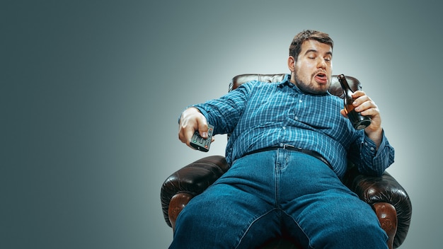 Retrato de hombre gordo caucásico con jeanse y whirt sentado en un sillón marrón aislado sobre fondo gris degradado. Ver televisión, beber cerveza y cambiar de canal, reír. Sobrepeso, despreocupado.