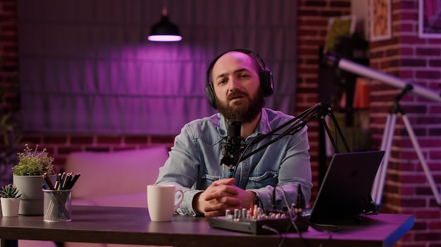 Retrato de un hombre grabando podcast usando un mezclador de audio y una computadora portátil hablando con un micrófono profesional en un estudio casero. Presentador de radio en línea con auriculares bebiendo café o té mientras transmite.