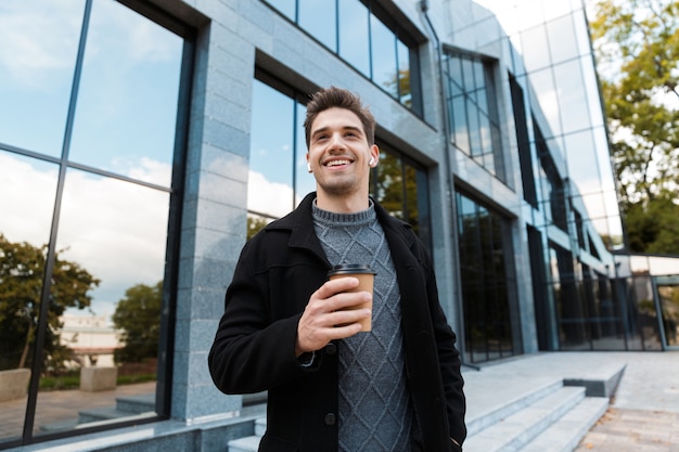 Retrato de hombre guapo de 30 años con audífonos sosteniendo una taza de papel de café para llevar mientras pasea frente al edificio de vidrio