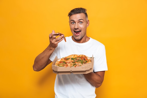 Retrato de hombre guapo de 30 años en camiseta blanca sosteniendo y comiendo pizza mientras está de pie aislado en amarillo