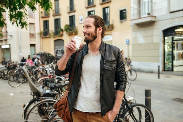 Retrato de hombre guapo de 30 años con chaqueta de cuero bebiendo café para llevar durante el paseo por las calles de la ciudad cerca de un montón de bicicletas