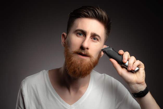 Retrato de un hombre guapo afeitando su barba con una afeitadora eléctrica por la mañana contra un fondo gris