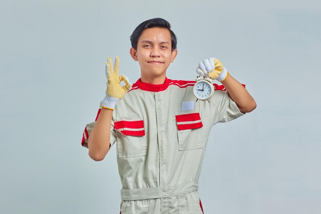 Retrato de hombre guapo alegre vestido con uniforme de mecánico sosteniendo el despertador y mostrando signo bien sobre fondo gris