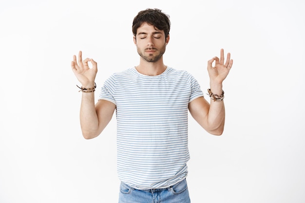 Retrato de hombre guapo, apuesto y guapo levantando las manos en gesto zen con los ojos cerrados, posando en posición de loto como haciendo práctica de yoga, liberando estrés, siendo tranquilo.