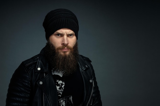 Retrato de hombre guapo con barba en chaqueta de cuero y sombrero mirando a la cámara