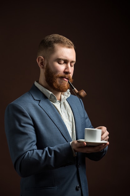 Retrato de hombre guapo con barba de jengibre sosteniendo una taza de café mientras fuma pipa