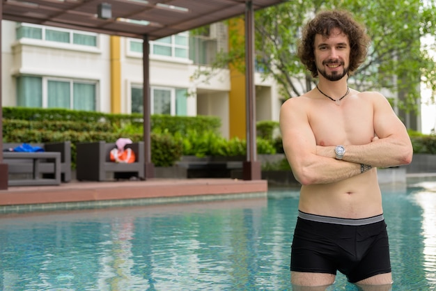 Retrato de hombre guapo con barba y pelo rizado descamisado relajante junto a la piscina en la ciudad de Bangkok, Tailandia