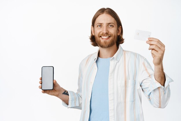 Retrato de hombre guapo con barba que muestra la tarjeta de crédito en la mano levantada, sosteniendo el teléfono móvil con interfaz de la aplicación, concepto de compras en línea, de pie en blanco