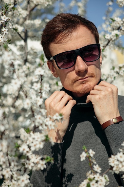 Retrato de hombre guapo en cuello alto y gafas de sol de pie entre árboles de flores en el parque
