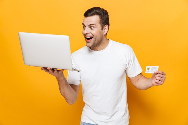 Retrato de un hombre guapo feliz con camiseta que se encuentran aisladas sobre la pared amarilla, mostrando la tarjeta de crédito