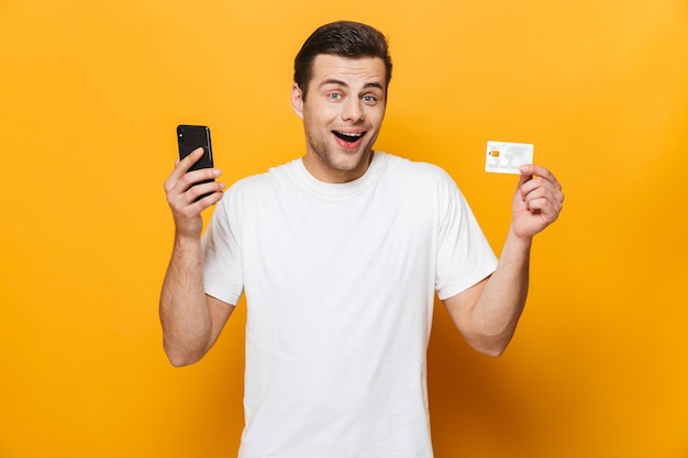 Retrato de un hombre guapo feliz con camiseta que se encuentran aisladas sobre la pared amarilla, mediante teléfono móvil, mostrando la tarjeta de crédito